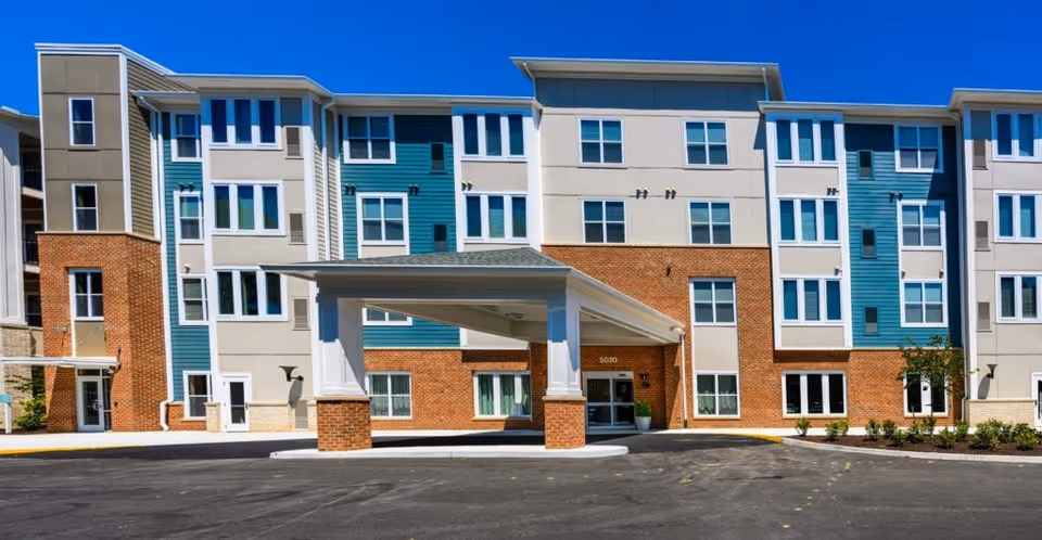 Exterior view of a modern multi-story senior living facility building with a covered entrance and a paved driveway under a clear blue sky.