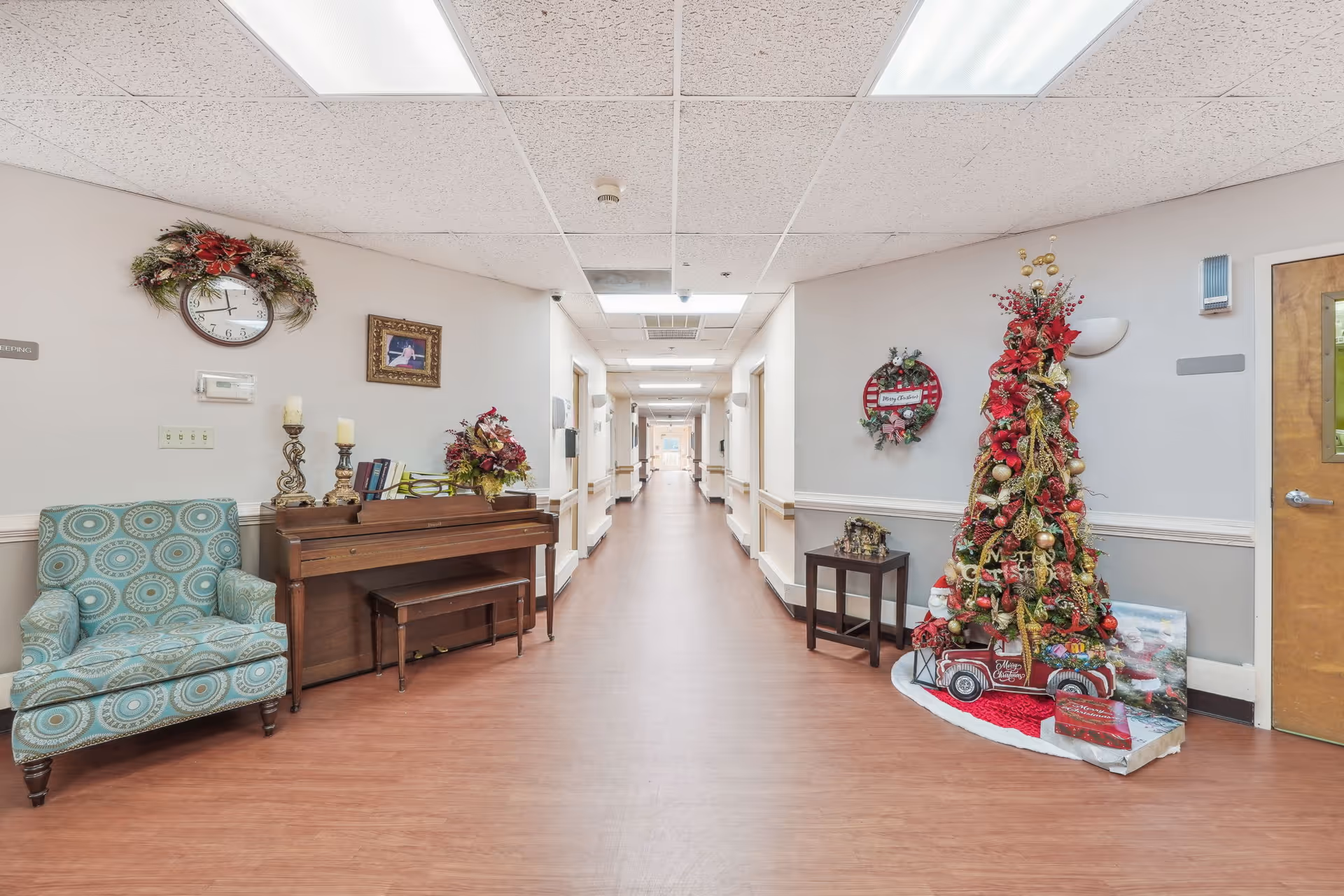 A long hallway in a senior living facility decorated for Christmas with a Christmas tree adorned with red and gold ornaments, a small red truck decoration, and a wreath on the wall. There is a wooden piano with candles and floral arrangements on top, a patterned armchair, and a small table with a nativity scene. The hallway has wood flooring, white walls with a chair rail, and fluorescent ceiling lights.