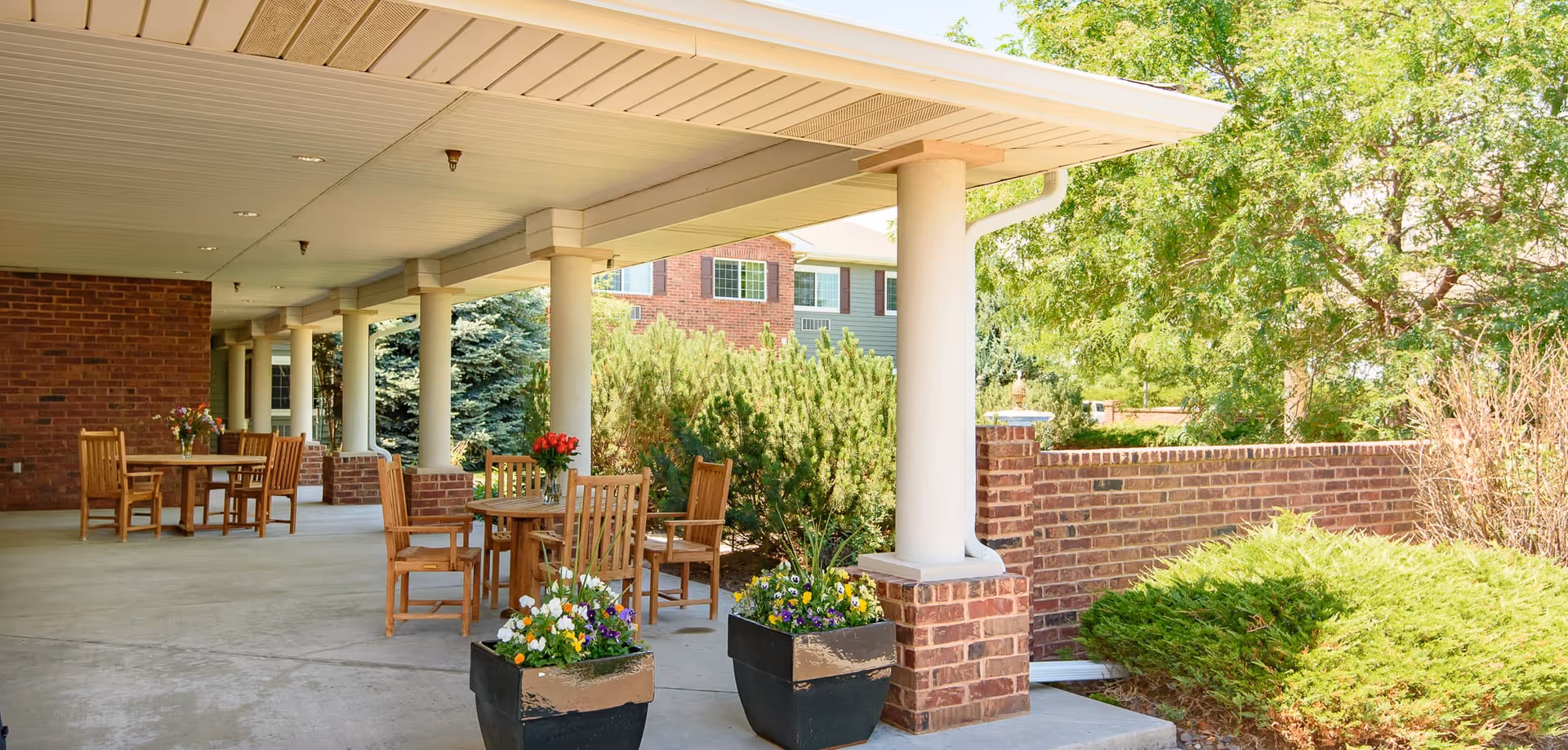 Covered outdoor patio with wooden tables and chairs, brick columns and walls, and potted flowers beside greenery.