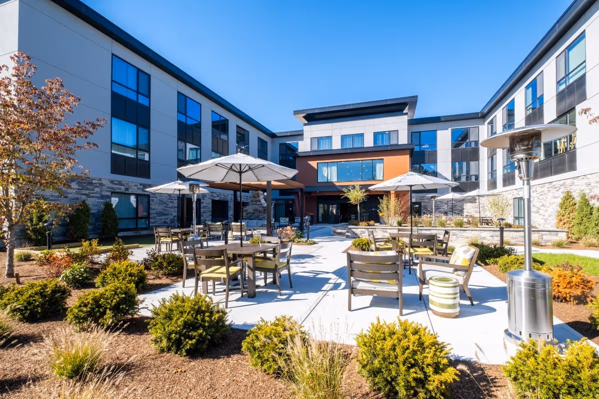 Outdoor courtyard area of Benchmark at Hanover featuring multiple tables with chairs and umbrellas, surrounded by landscaped bushes and trees, with a modern three-story building in the background under a clear blue sky.
