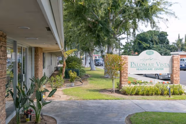 Outdoor view of Palomar Vista Healthcare Center showing the entrance area with a sidewalk, plants, trees, and a sign displaying the facility's name and address number 201.