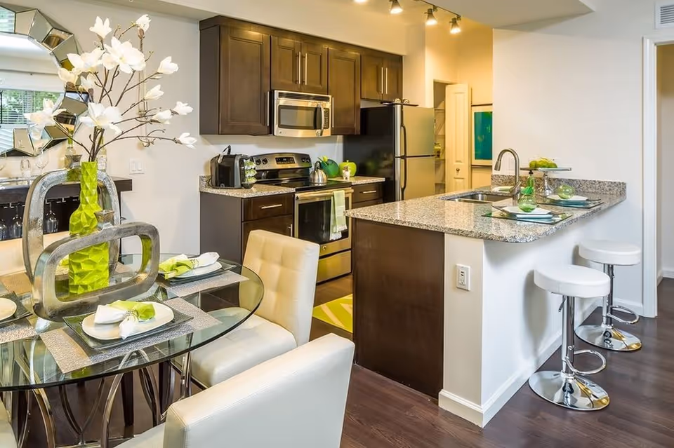 Modern kitchen and dining area with dark wood cabinets, stainless steel appliances, a granite countertop island with two white bar stools, and a glass dining table set with white cushioned chairs and green decorative accents including a vase with white flowers.