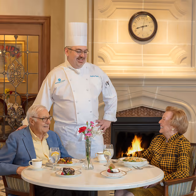 A chef stands and smiles while talking with two elderly residents seated at a dining table set with plates, drinks, and a vase of flowers in front of a lit fireplace.