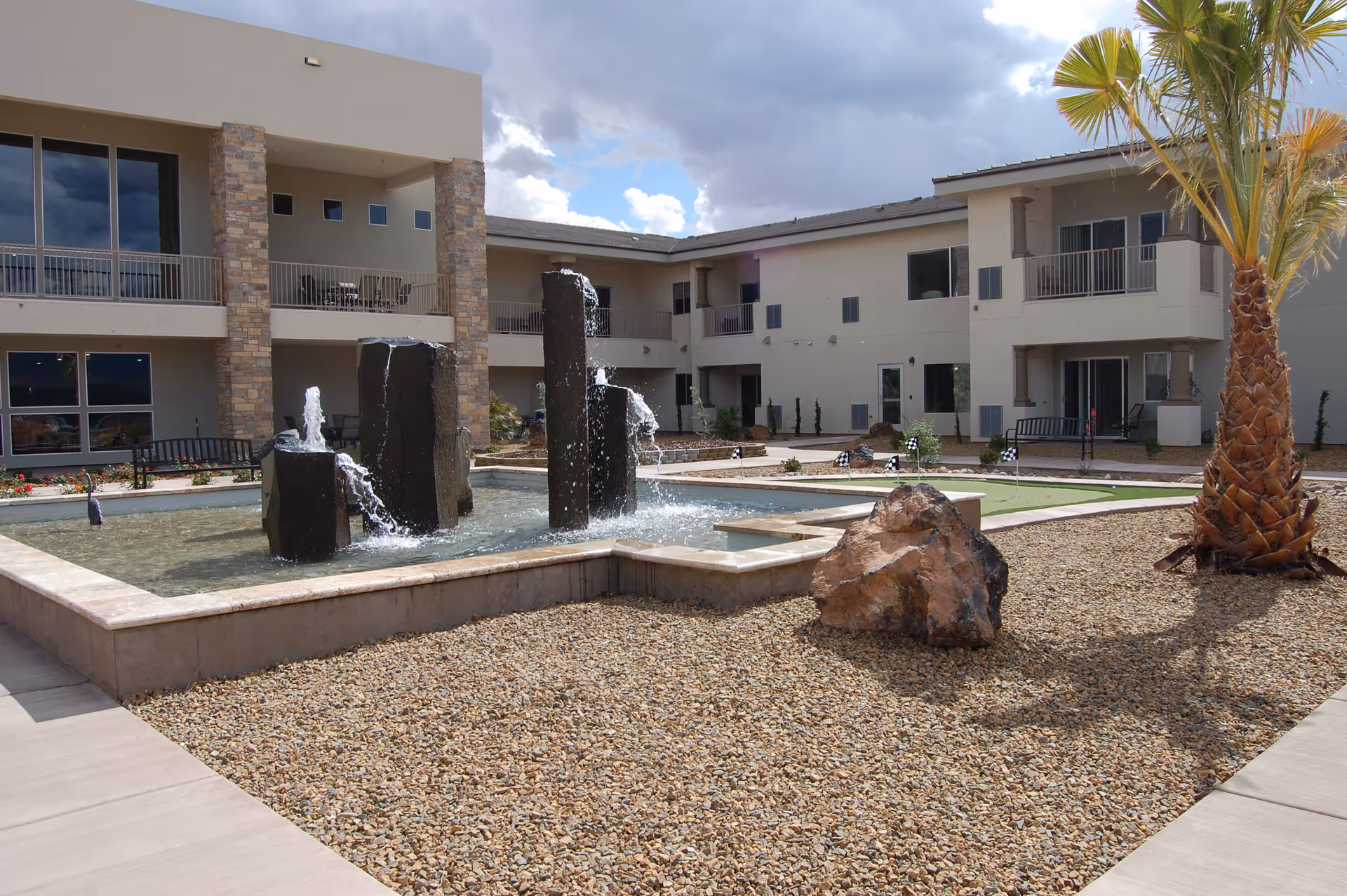 Outdoor courtyard area of a senior living facility featuring a water fountain with multiple vertical stone pillars spraying water, surrounded by gravel landscaping, a palm tree, and a large rock. The building has two stories with balconies and large windows overlooking the courtyard.