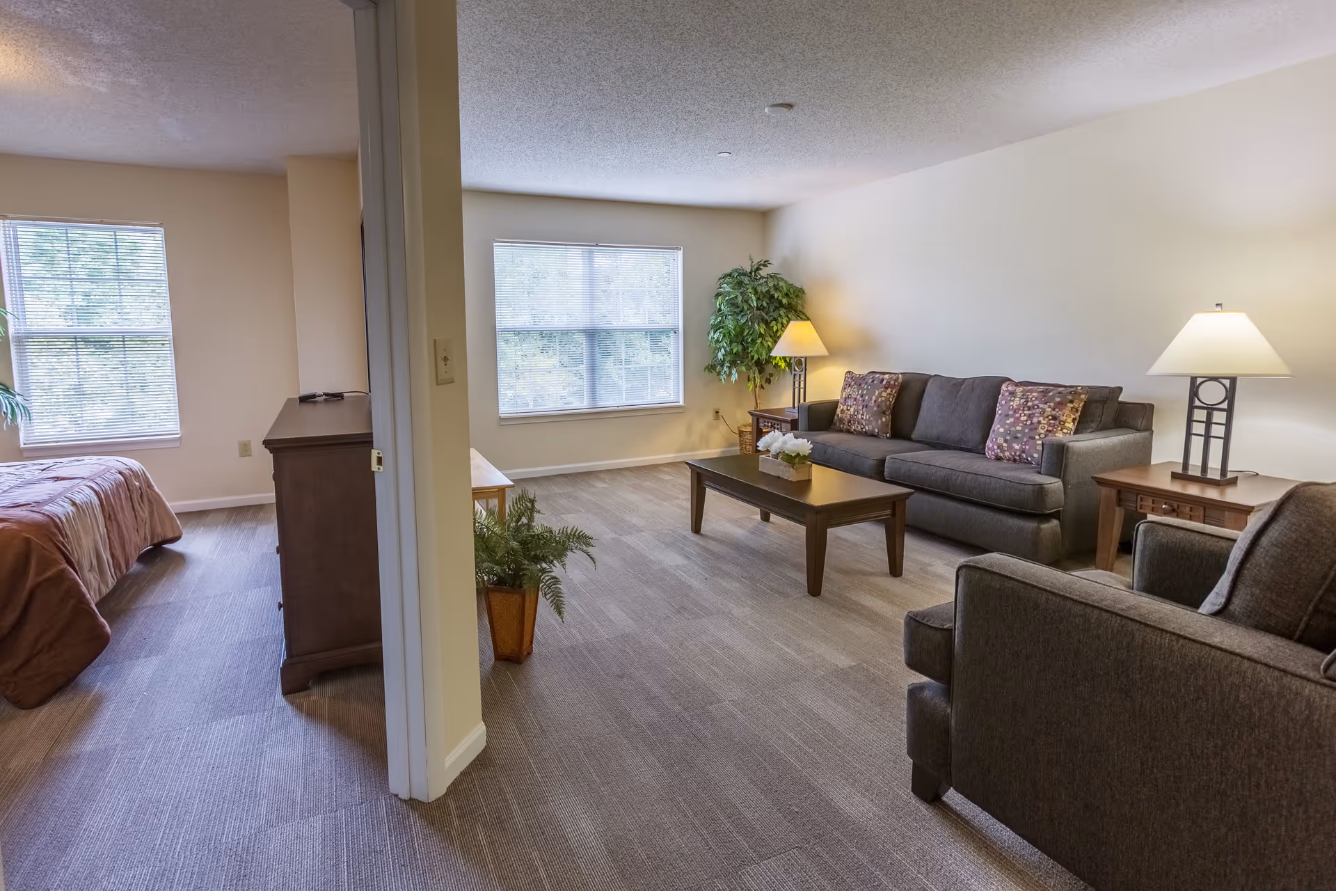 Interior view of a senior living facility apartment showing a living room and a bedroom. The living room has a gray sofa with patterned pillows, a matching armchair, two wooden side tables with lamps, a wooden coffee table with a small plant, and a potted plant in the corner. The bedroom has a bed with a brown bedspread, a wooden dresser, and two windows letting in natural light.