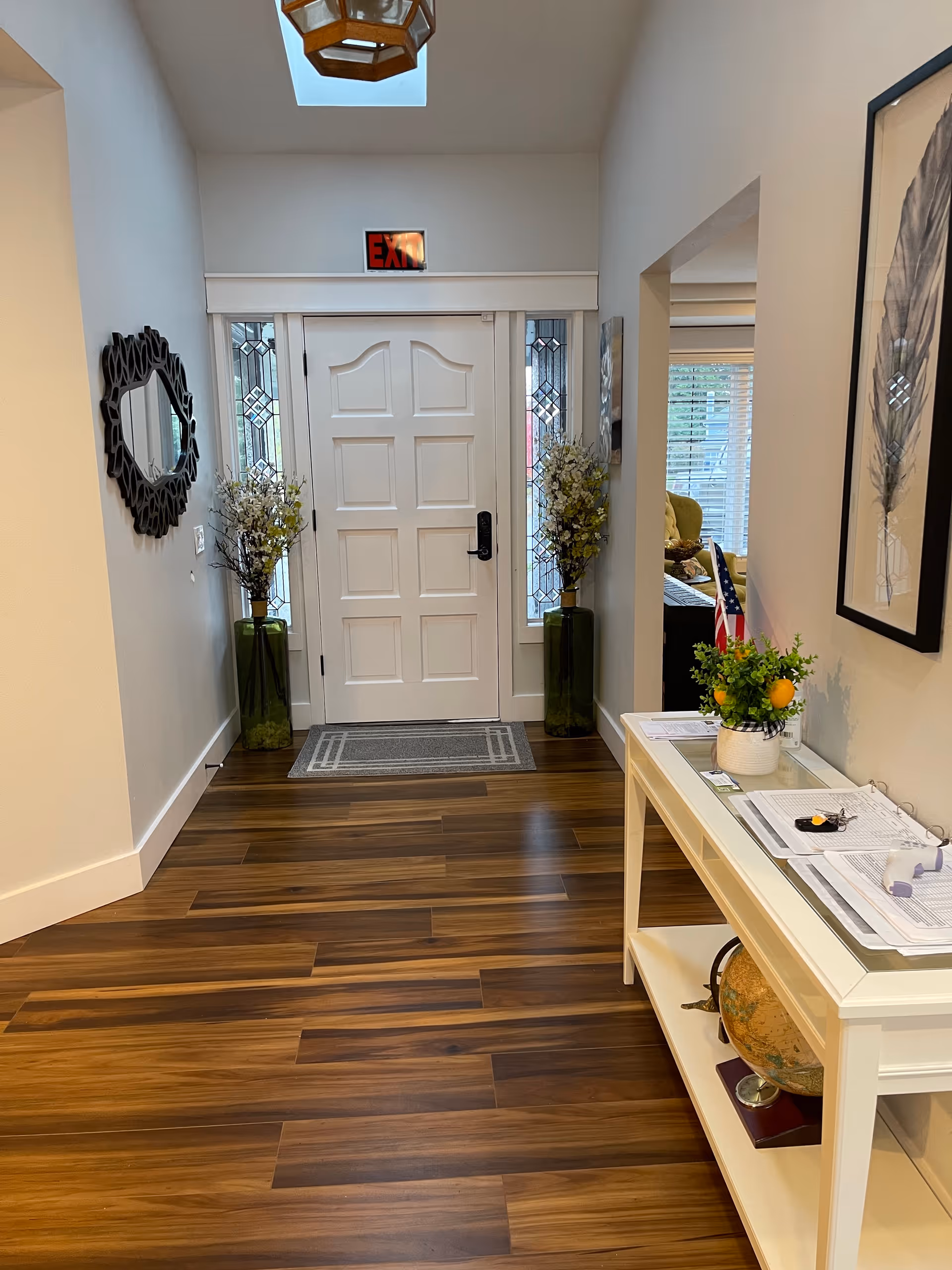 Entryway of a facility with a white front door featuring decorative glass panels on each side. Two tall green vases with white flowers are placed on either side of the door. A rectangular gray doormat is in front of the door on a wooden floor. On the right side, there is a white console table with papers, a small plant, and a globe on the lower shelf. A framed artwork of feathers hangs on the wall above the table. A decorative round mirror is mounted on the left wall. The ceiling has a skylight and a hanging light fixture. An illuminated red EXIT sign is above the door.