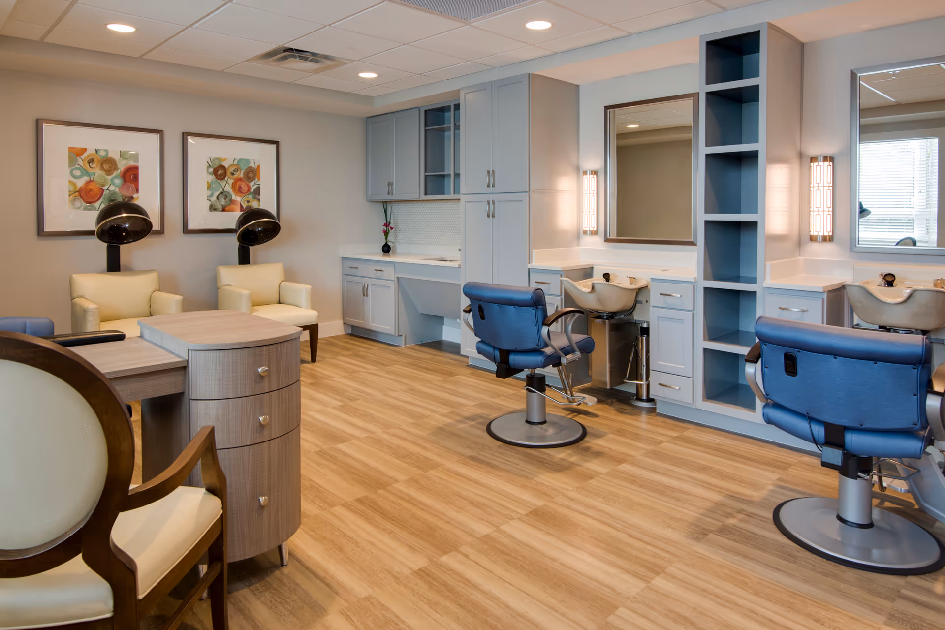 Interior of a salon area in a senior living facility with two blue salon chairs in front of sinks and mirrors, two beige armchairs with black hair dryers behind them, light wood flooring, light gray cabinetry, and colorful framed artwork on the wall.