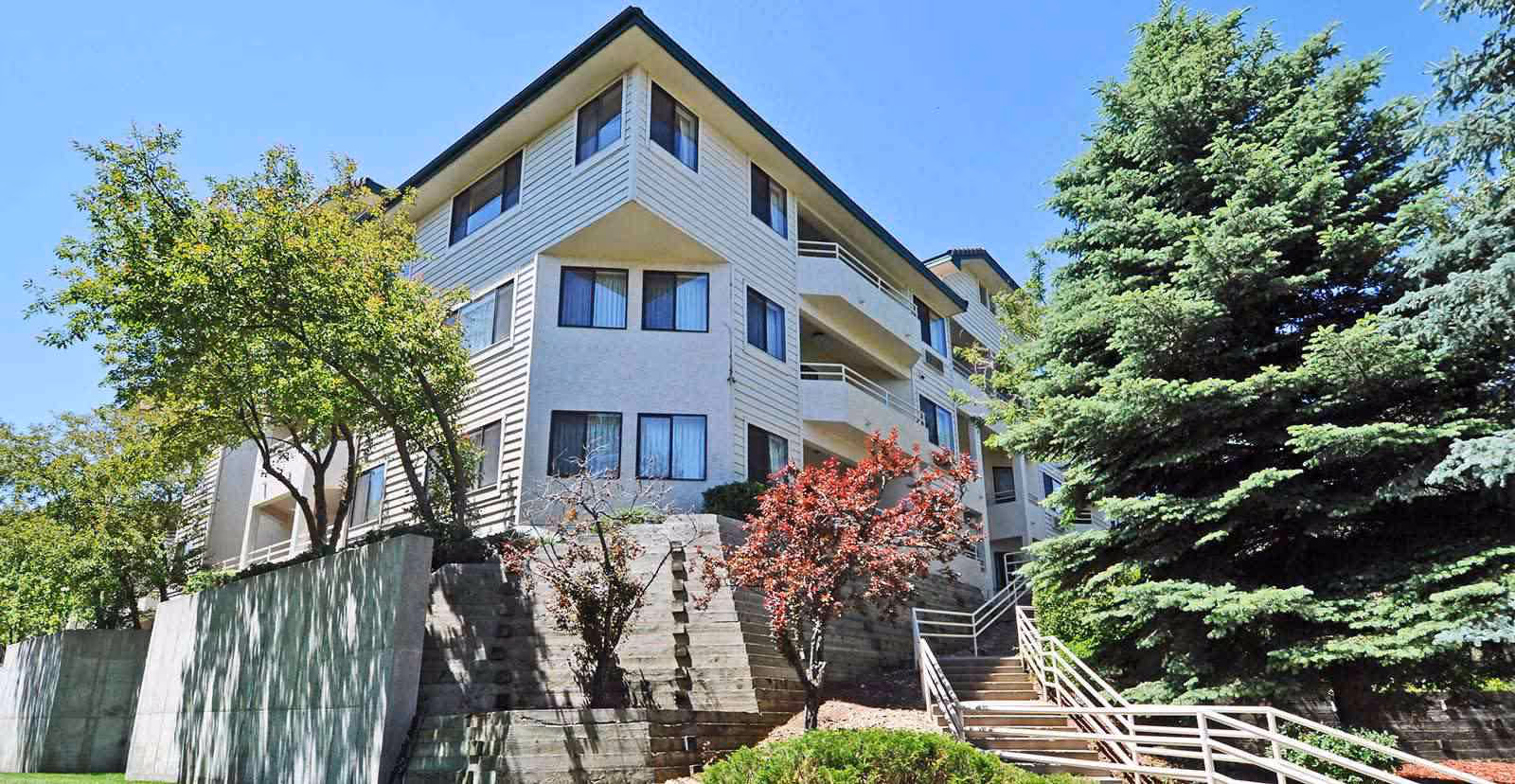 Three-story light-colored apartment building with balconies, an exterior stairway, and landscaped trees under a clear blue sky.