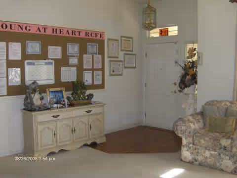 Reception/sitting area with a bulletin board reading 'Young at Heart RCFE', a console table, floral sofa, and a door with an EXIT sign.