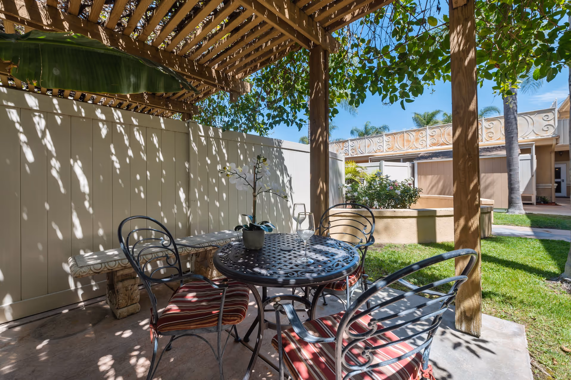 Outdoor patio area with a round metal table and four metal chairs with red striped cushions under a wooden pergola with leafy vines providing shade. A potted orchid and a wine glass are on the table. The patio is adjacent to a beige fence and a garden area with green grass and plants.