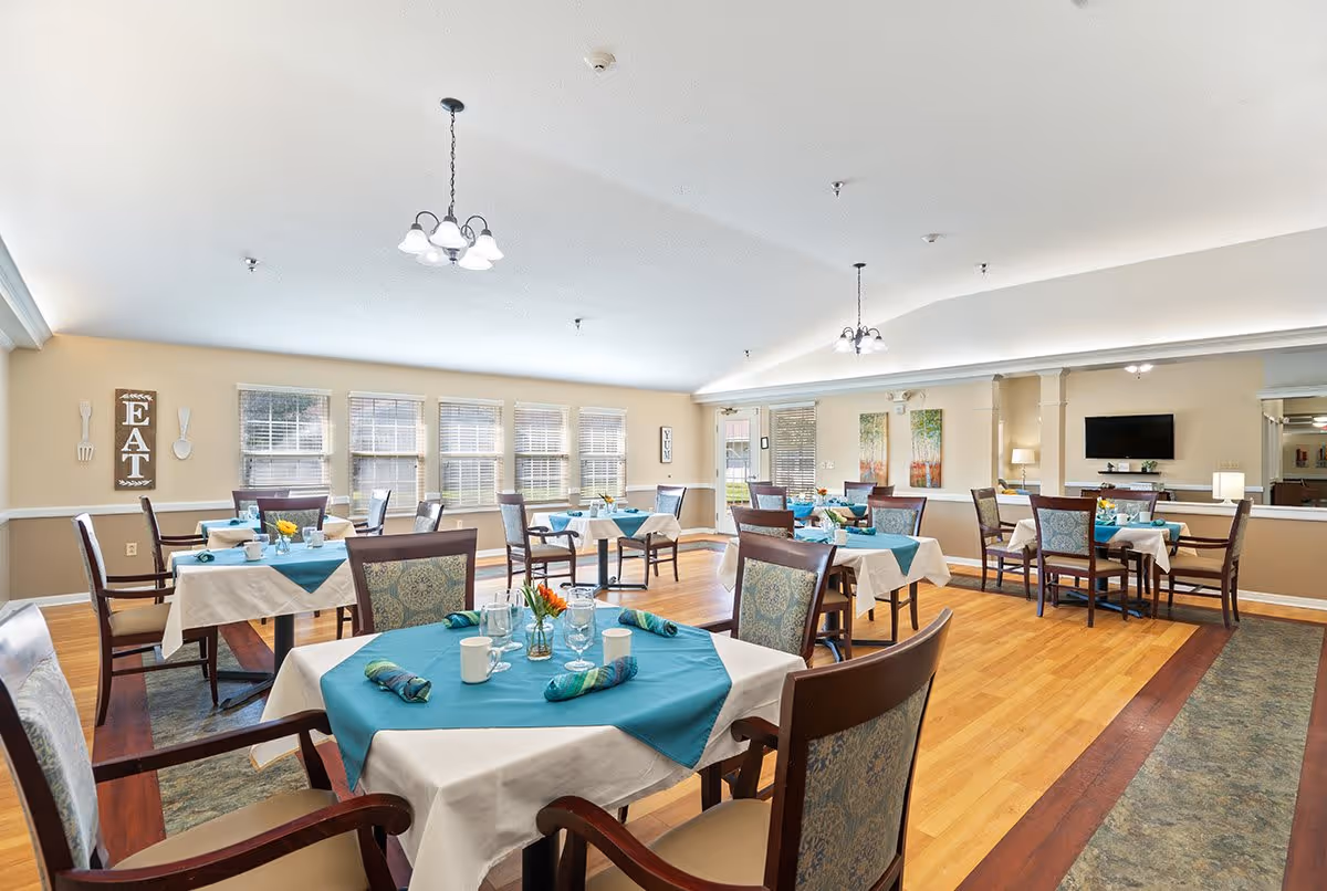 A bright dining room with several tables covered in white tablecloths and teal table runners. Each table is set with glasses, cups, and napkins, and some have small flower arrangements. The room has large windows letting in natural light, beige walls, and wooden flooring. There are decorative signs on the walls, including one that says 'EAT'.