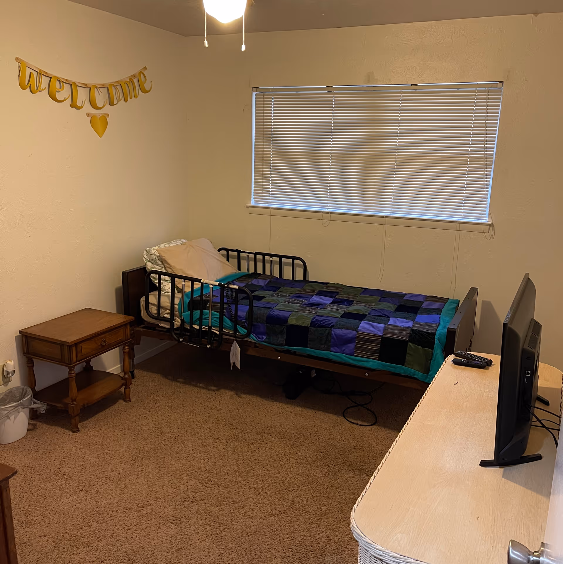 Small bedroom with a single bed with black safety rails and a patchwork blanket, a nightstand, window blinds, a TV on a dresser, and a 'welcome' banner on the wall.