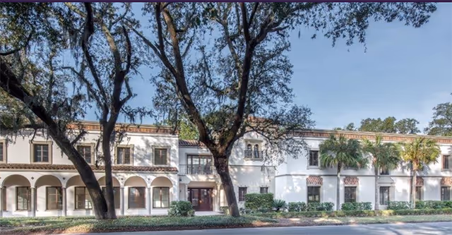 White two-story Mediterranean-style building front with arched colonnade, palm and oak trees lining the street.