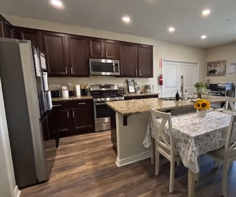Open kitchen with dark wood cabinets, stainless steel appliances, a granite island, and a small dining table with a floral tablecloth and sunflowers.