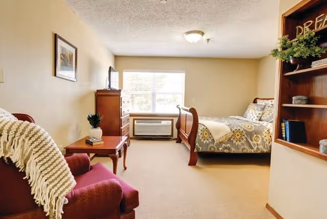 A cozy bedroom in a senior living facility with a wooden bed featuring patterned bedding, a wooden dresser with a TV on top, a red armchair with a white throw blanket, a small wooden side table with a plant and books, a window with blinds, and a wooden bookshelf with decorative items.