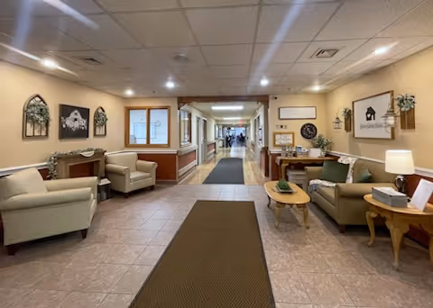 A well-lit hallway in a senior living facility with comfortable seating areas on both sides. The seating areas include armchairs and small tables with lamps and decorative items. The walls are adorned with framed pictures and wall decorations. The floor is tiled with a brown runner rug in the center leading down the hallway.