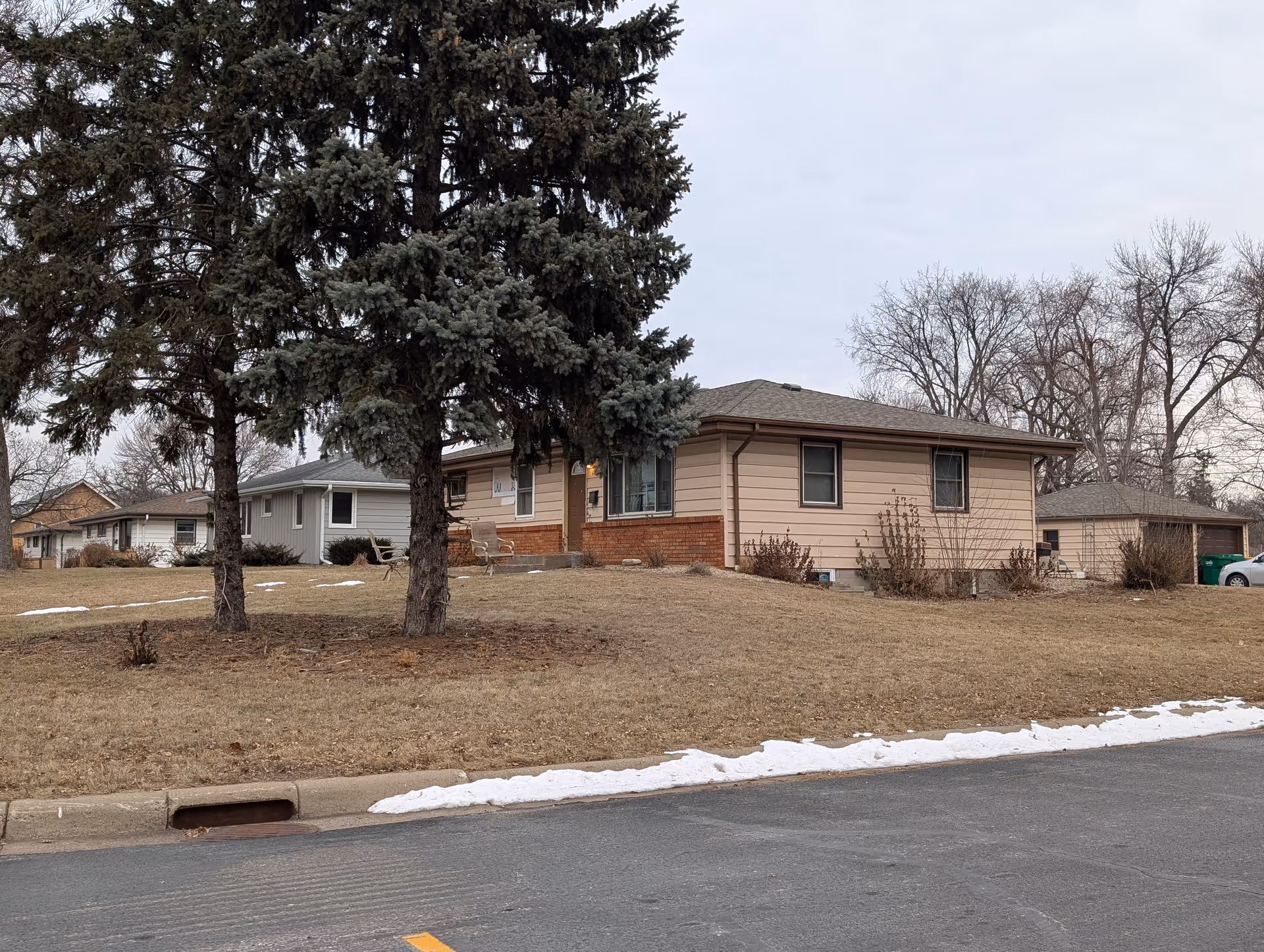 Exterior view of a single-story residential building with beige siding and a brick base, surrounded by leafless trees and a lawn with patches of snow. A paved road is in the foreground.