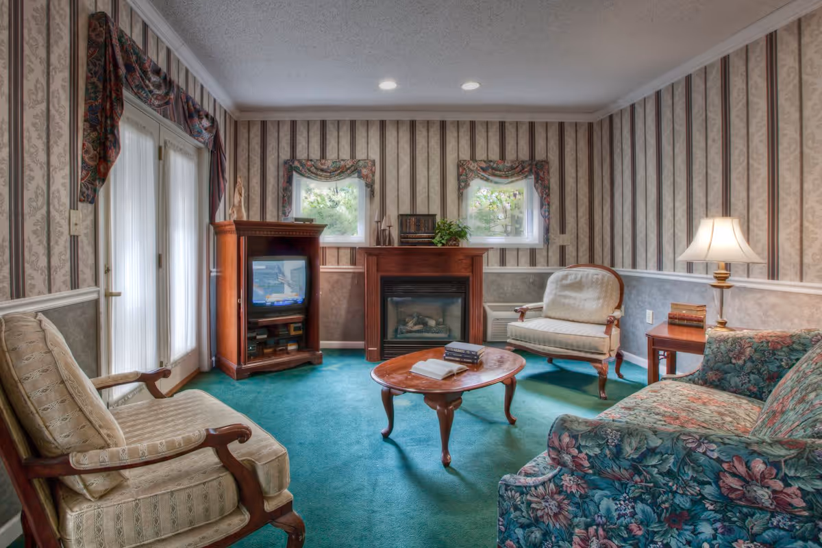 A cozy living room with vintage-style furniture including a floral patterned sofa, two armchairs, a wooden coffee table with books, a wooden TV cabinet with a television, and a fireplace. The room has green carpet, striped wallpaper with floral accents, two small windows with floral valances, and a table lamp on a side table.