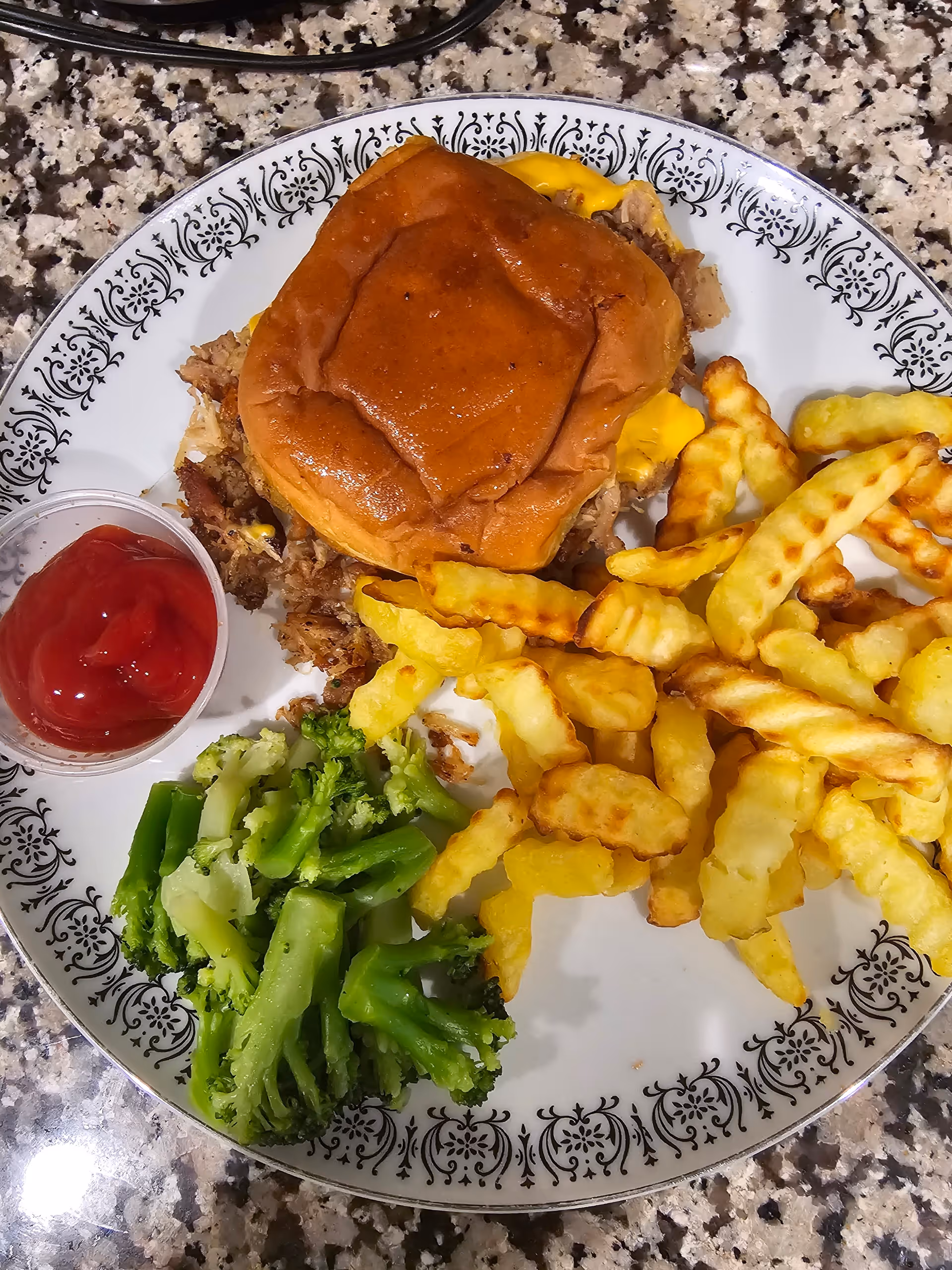 Decorative plate with a sandwich, crinkle-cut fries, steamed broccoli and a small cup of ketchup on a speckled countertop.