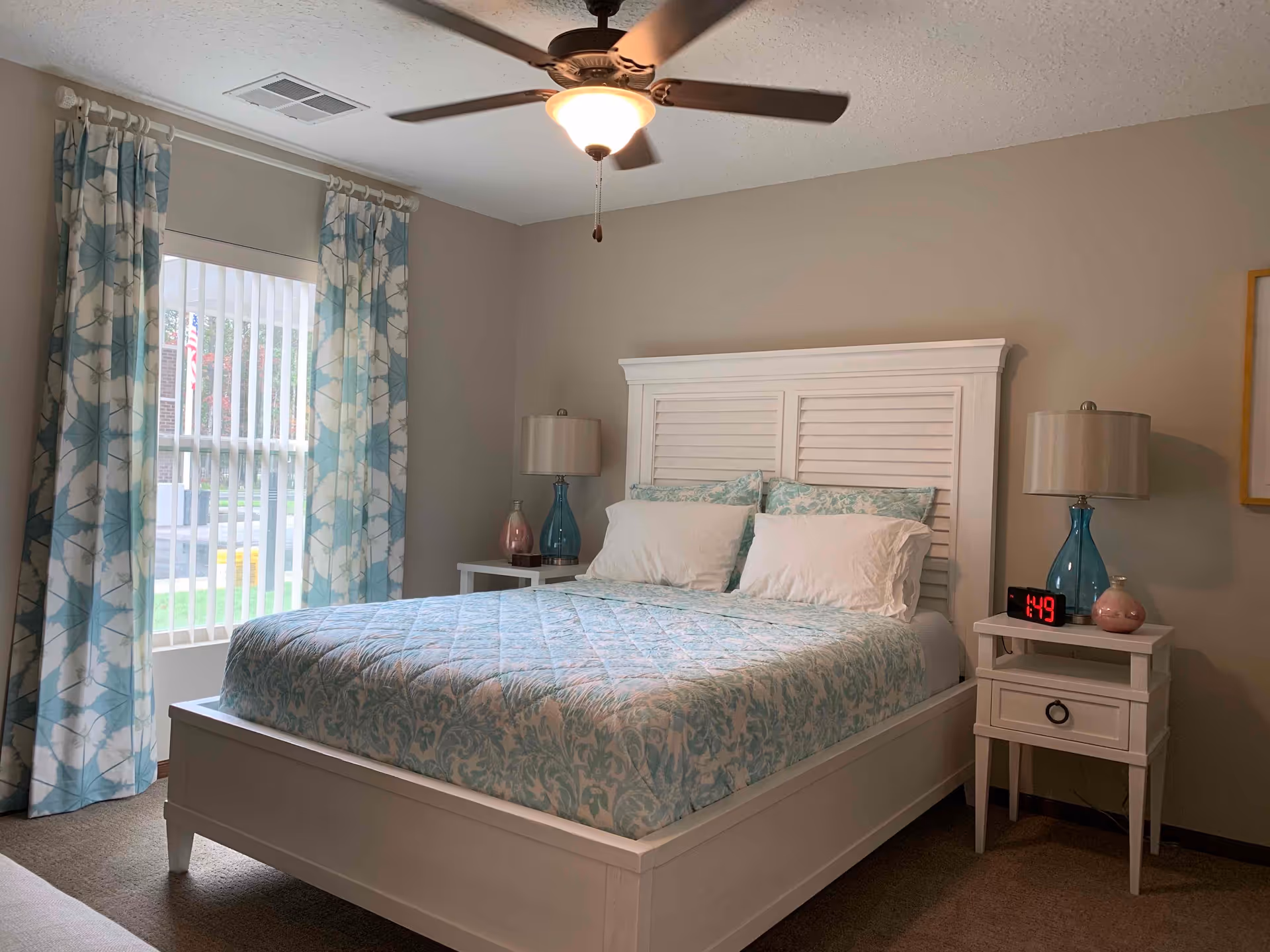 A bedroom with a white wooden bed frame and a blue patterned bedspread. There are two white pillows and two blue patterned pillows on the bed. On each side of the bed, there is a white nightstand with a blue lamp and decorative items. A digital clock on the right nightstand shows the time 1:49. The room has beige walls, a ceiling fan with a light, and a window with vertical blinds and blue floral curtains.