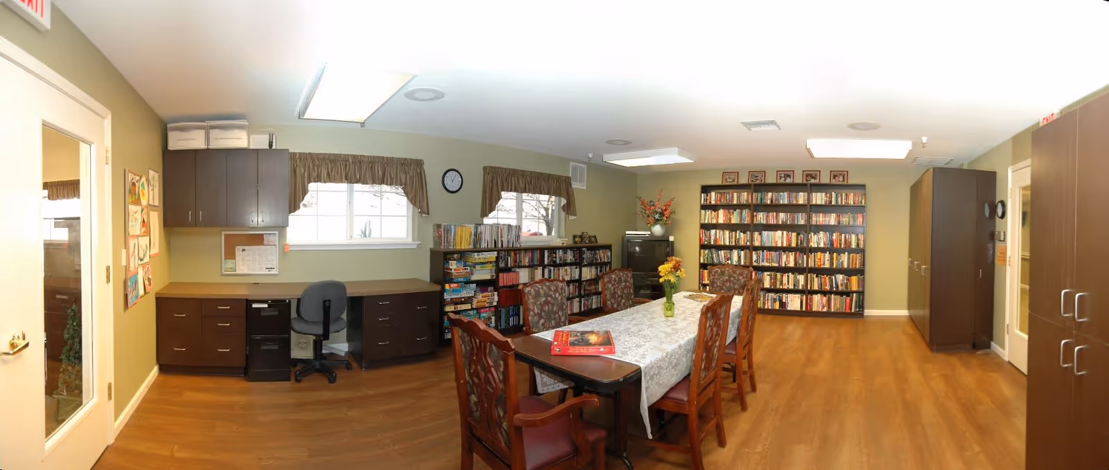 A well-lit room with wooden flooring featuring a long table covered with a white lace tablecloth and surrounded by six wooden chairs with patterned cushions. Against the walls are multiple bookshelves filled with books and board games. There is a desk with a black office chair under two windows with brown valances. The walls are painted light green, and there are cabinets and a clock on the wall. A vase with flowers is placed on the table.