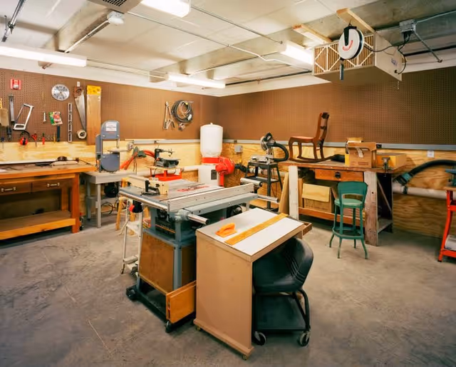 A woodworking workshop with various tools and equipment including a table saw, workbenches, clamps, and chairs. The walls have pegboards with hanging tools and supplies. The floor is concrete and the room is well-lit with fluorescent lights.