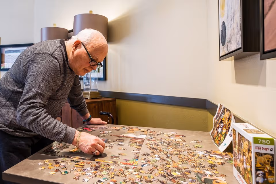 An elderly man wearing glasses and a striped sweater is leaning over a table working on a jigsaw puzzle in a well-lit room with framed pictures on the wall and a lamp on a wooden cabinet in the background.