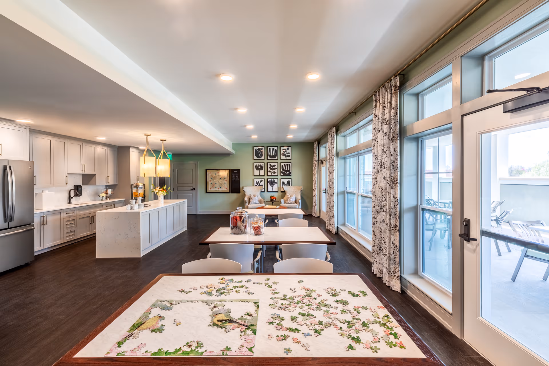 A bright and spacious common area in a senior living facility featuring a modern kitchen with white cabinetry and stainless steel appliances on the left, a seating area with two cushioned chairs and framed artwork on the far wall, and large windows with patterned curtains on the right letting in natural light. In the foreground, there are tables with chairs, one of which has a partially completed jigsaw puzzle. A door leads to an outdoor patio with additional seating.