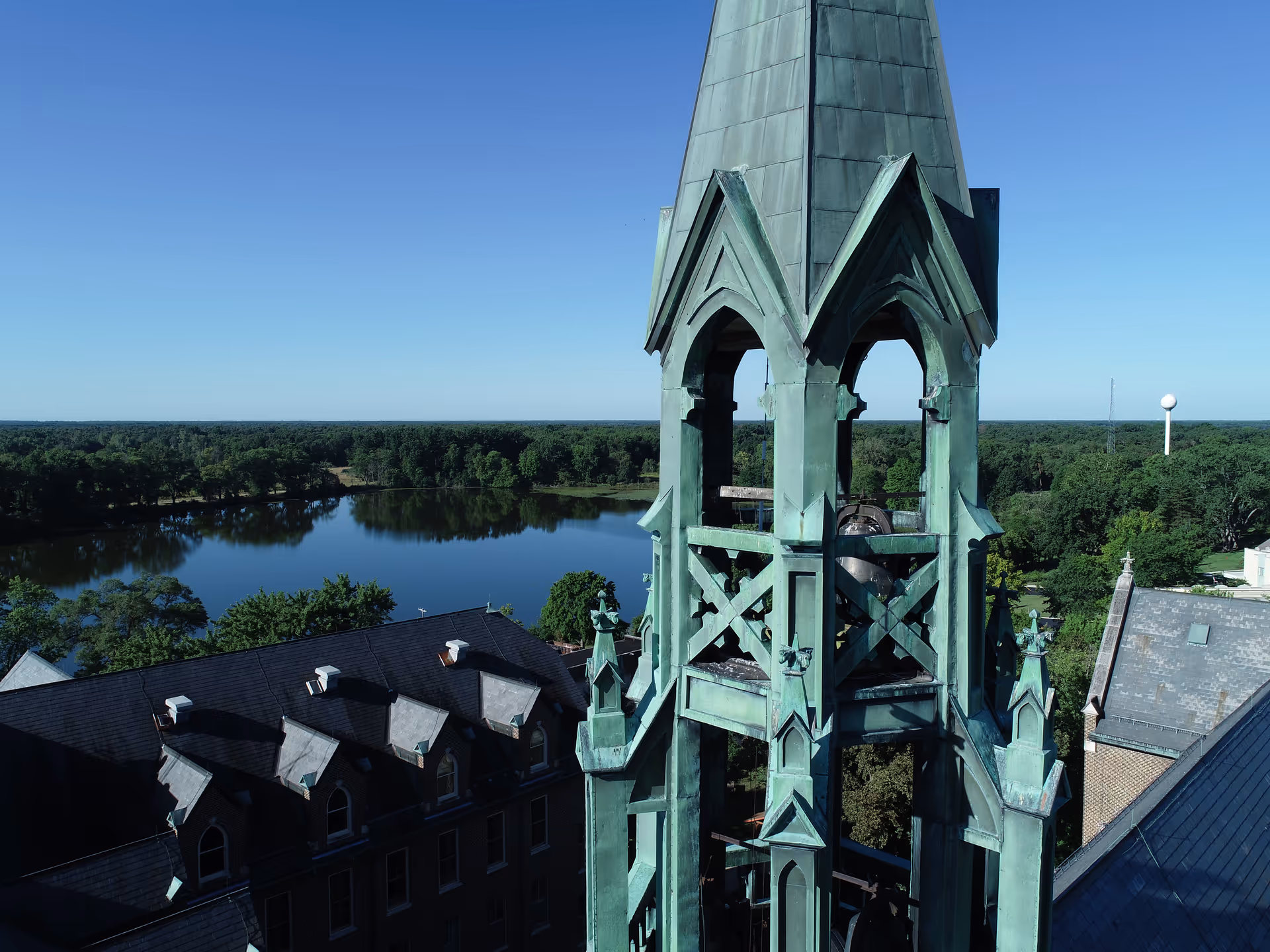 Aerial view of a tall, greenish copper bell tower with a large bell inside, part of a historic building with a dark roof. In the background, there is a calm lake surrounded by trees under a clear blue sky.