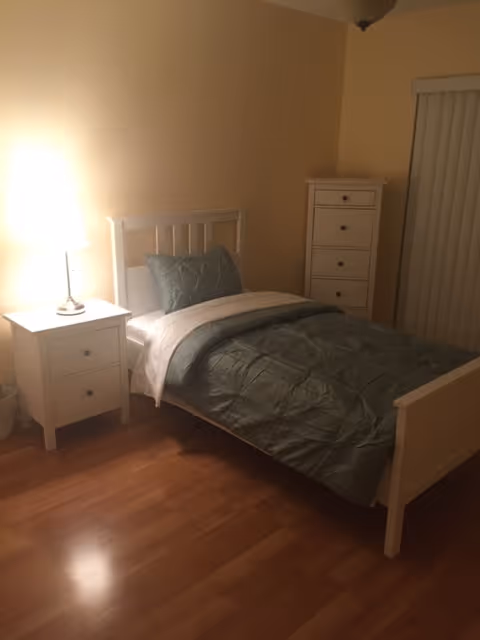 A small bedroom with a single white bed made with green bedding and a white blanket. Next to the bed is a white nightstand with a lamp on it. There is a tall white dresser in the corner and vertical blinds covering a window or door. The room has wooden flooring and beige walls.