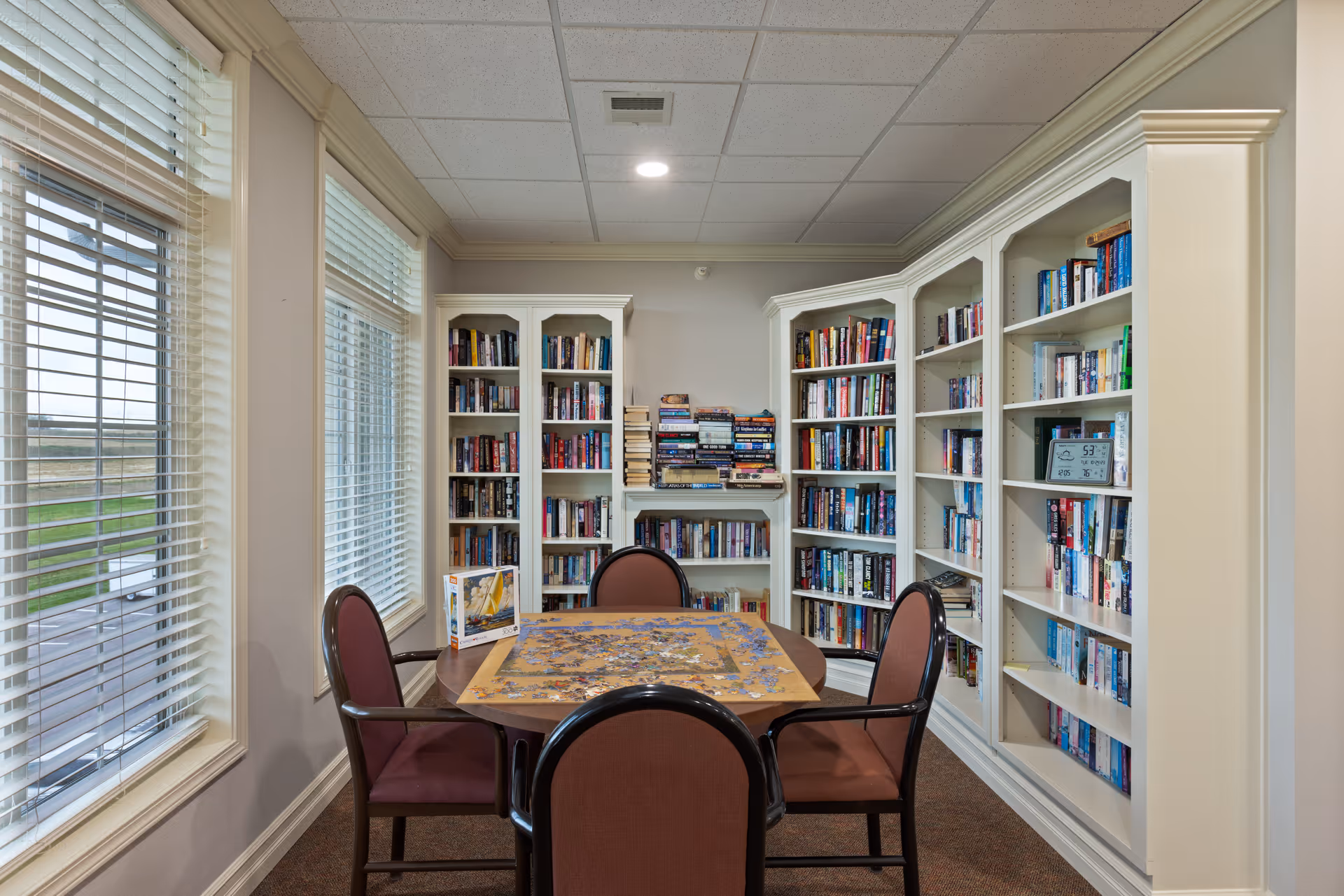 Small senior living reading room with bookshelves along the walls, a table holding a jigsaw puzzle and four chairs, and large windows with blinds.