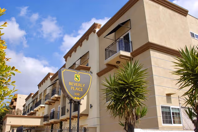 Exterior view of Silverado Beverly Place Memory Care Community building with beige walls, multiple balconies, and a sign in front that reads 'Beverly Place 330' under a blue sky with some clouds.