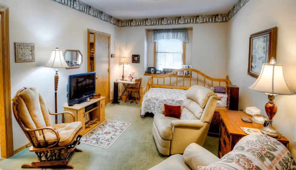 A cozy bedroom with a wooden bed covered in a white floral bedspread, a beige recliner chair with a red pillow, a wooden rocking chair, a TV on a wooden stand, a small desk with a chair and lamp near the window, and various lamps and framed pictures on the walls.