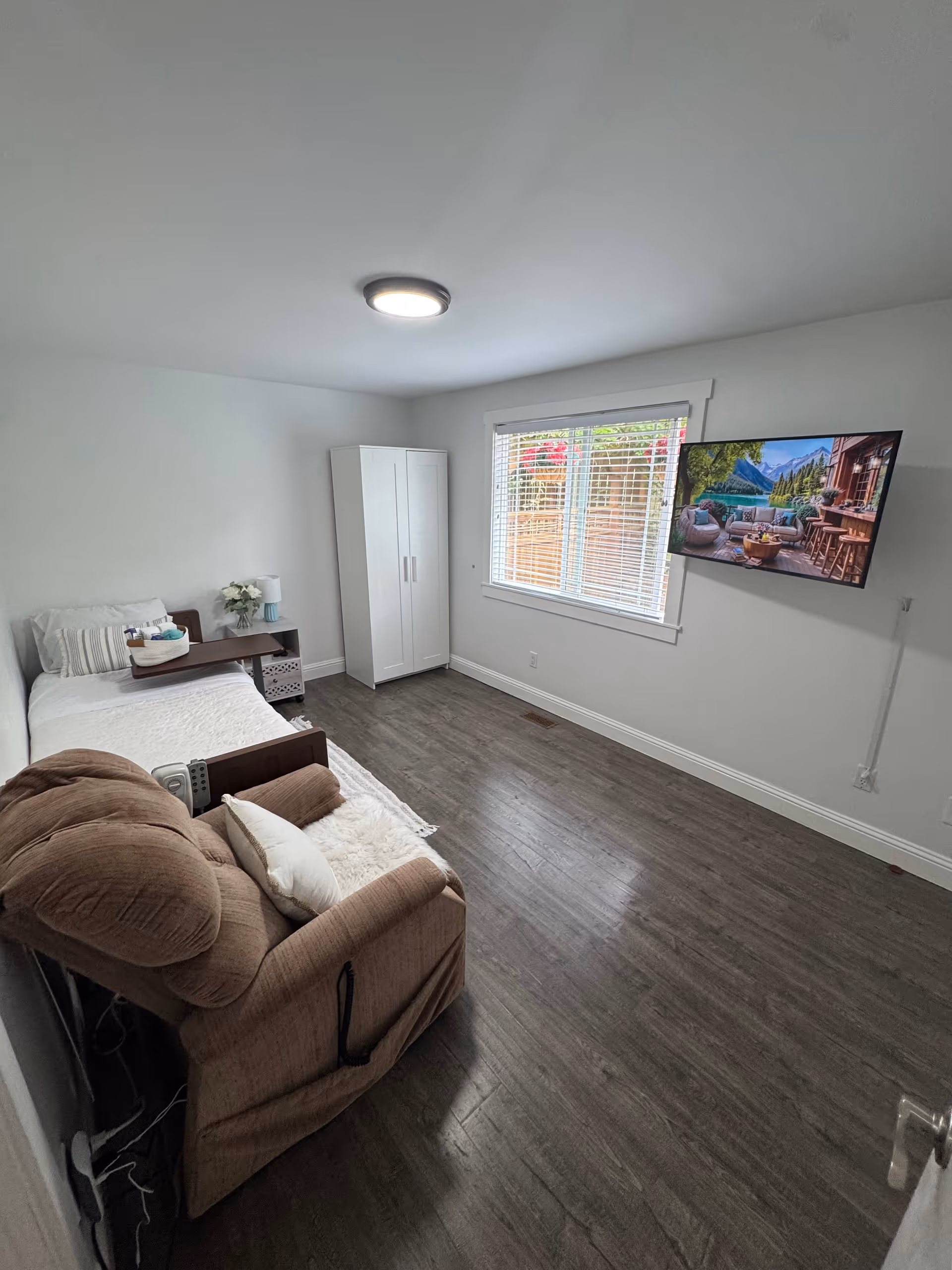 A small, clean room with a single bed covered in white bedding, a brown recliner chair with a white pillow, a white wardrobe, a small bedside table with a lamp and flowers, and a wall-mounted flat-screen TV displaying an outdoor patio scene. The room has wooden flooring, white walls, and a window with blinds letting in natural light.