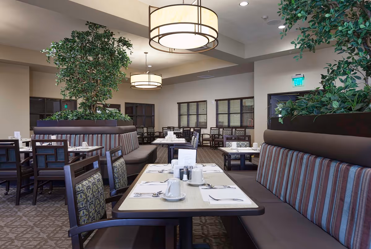 A spacious dining room in a senior living facility with tables set for meals. The room features cushioned chairs and booth seating with striped upholstery. Large potted plants are placed on top of the booth dividers, and modern circular pendant lights hang from the ceiling. Windows with blinds line the walls, and the overall decor is warm and inviting.