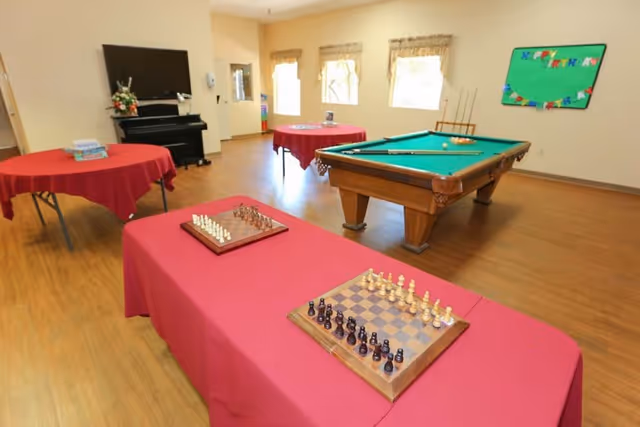 A recreational room with a pool table, two chess boards on a table covered with a red cloth, and two additional round tables also covered with red cloth. The room has wooden flooring, three windows with curtains, a piano with a TV mounted above it, and a colorful 'Happy Birthday' sign on the wall.