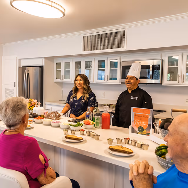 A chef and a staff member smile behind a kitchen island while senior residents sit across with plates and cooking ingredients laid out.