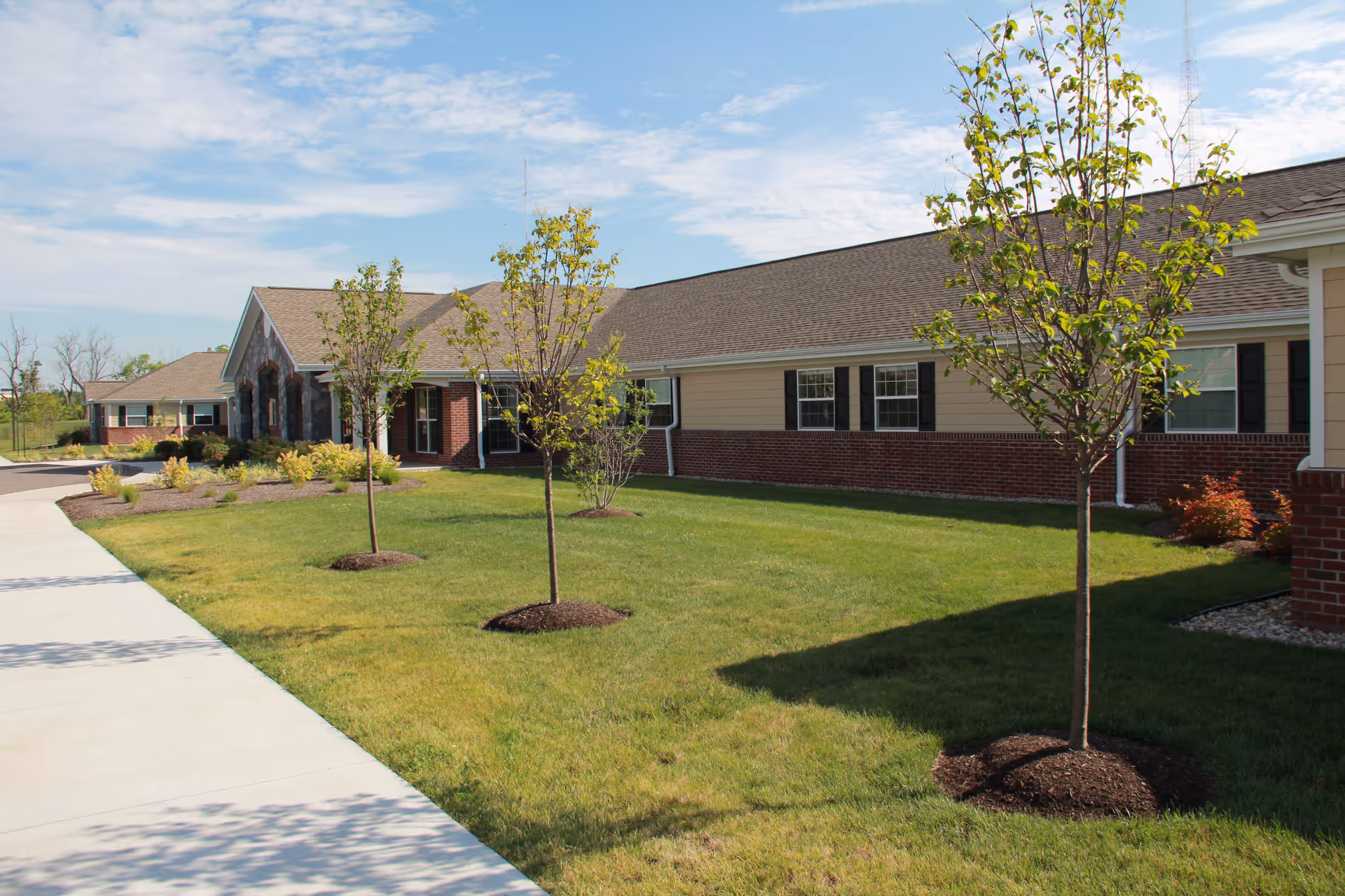 Exterior view of a single-story senior living facility building with a brick and beige siding facade, several young trees planted in a grassy lawn, and a concrete walkway leading to the entrance under a partly cloudy sky.