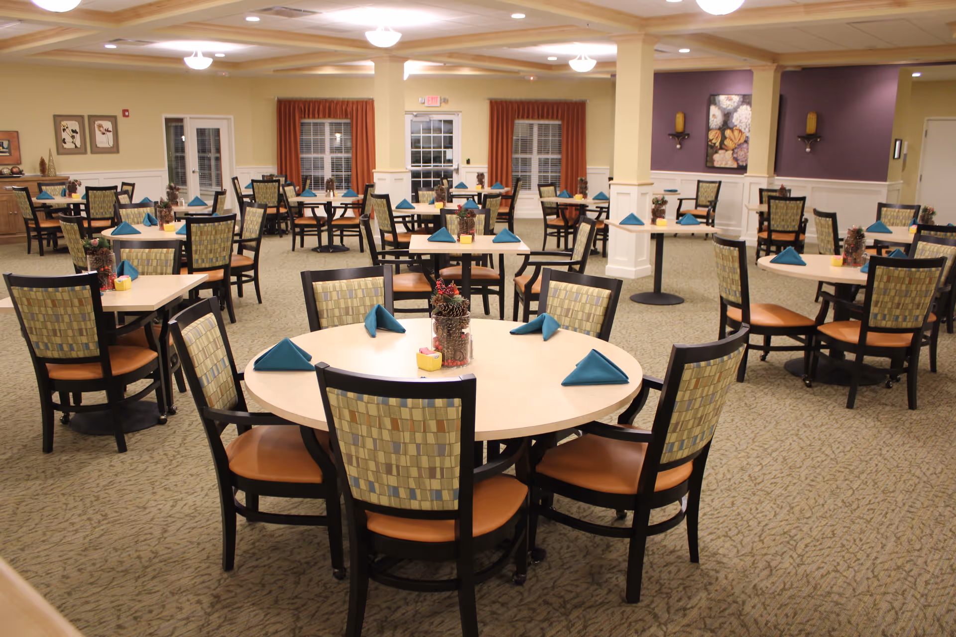 A spacious dining room with multiple round and square tables, each surrounded by chairs with patterned backs and brown seats. The tables are set with folded teal napkins and small decorative centerpieces. The room has beige walls with white wainscoting, carpeted floors, and ceiling lights. There are windows with red curtains and some wall decorations.