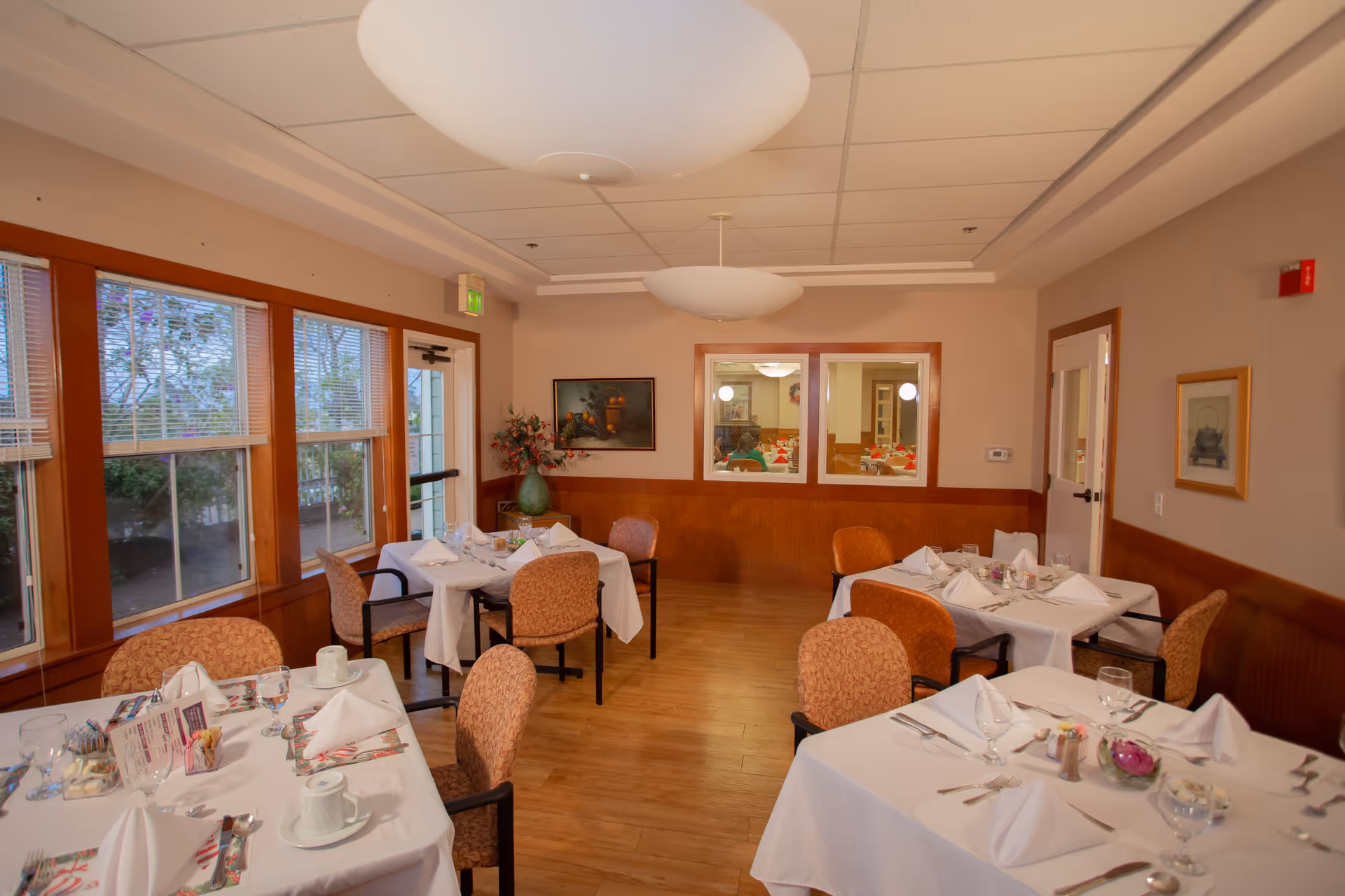 A dining room with several tables covered in white tablecloths, each set with folded napkins, glassware, and utensils. The room has wooden floors, beige walls with wood paneling, large windows on the left side, and two large ceiling light fixtures. There is a door and framed artwork on the walls.