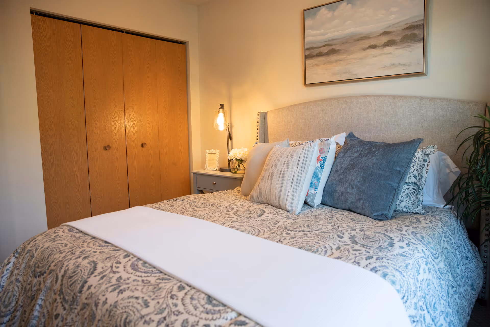 A cozy bedroom with a neatly made bed featuring a patterned bedspread and multiple decorative pillows. There is a wooden closet with double doors on the left side, a small bedside table with a lamp and flowers, and a framed landscape painting hanging on the wall above the bed.