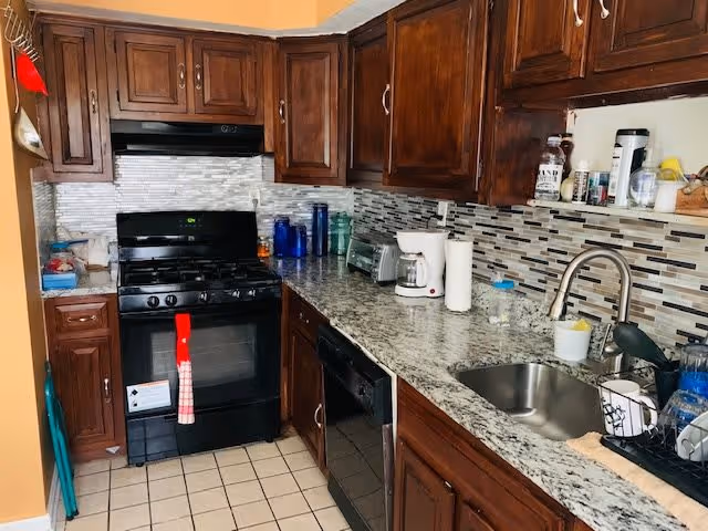 A kitchen with dark wooden cabinets, a black gas stove with a red and white towel hanging on the oven door, a granite countertop with various items including a coffee maker, paper towels, and cups, a stainless steel sink, and a tiled backsplash in shades of gray and white.