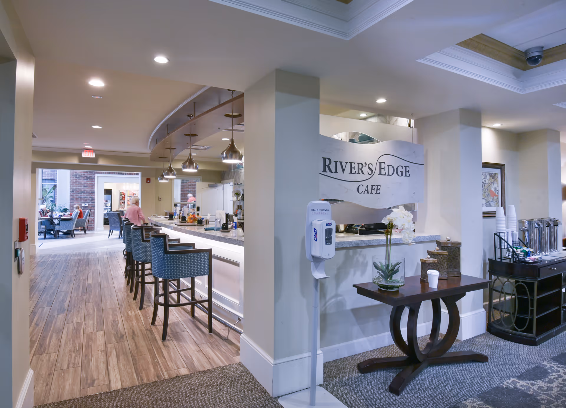 Interior view of River's Edge Cafe at Riverstone at Friendship Village of Dublin, featuring a long counter with bar stools, pendant lighting, a hand sanitizer dispenser, a small table with a flower arrangement, and a beverage station with cups and coffee urns. In the background, there is a seating area with tables and chairs and a person sitting.