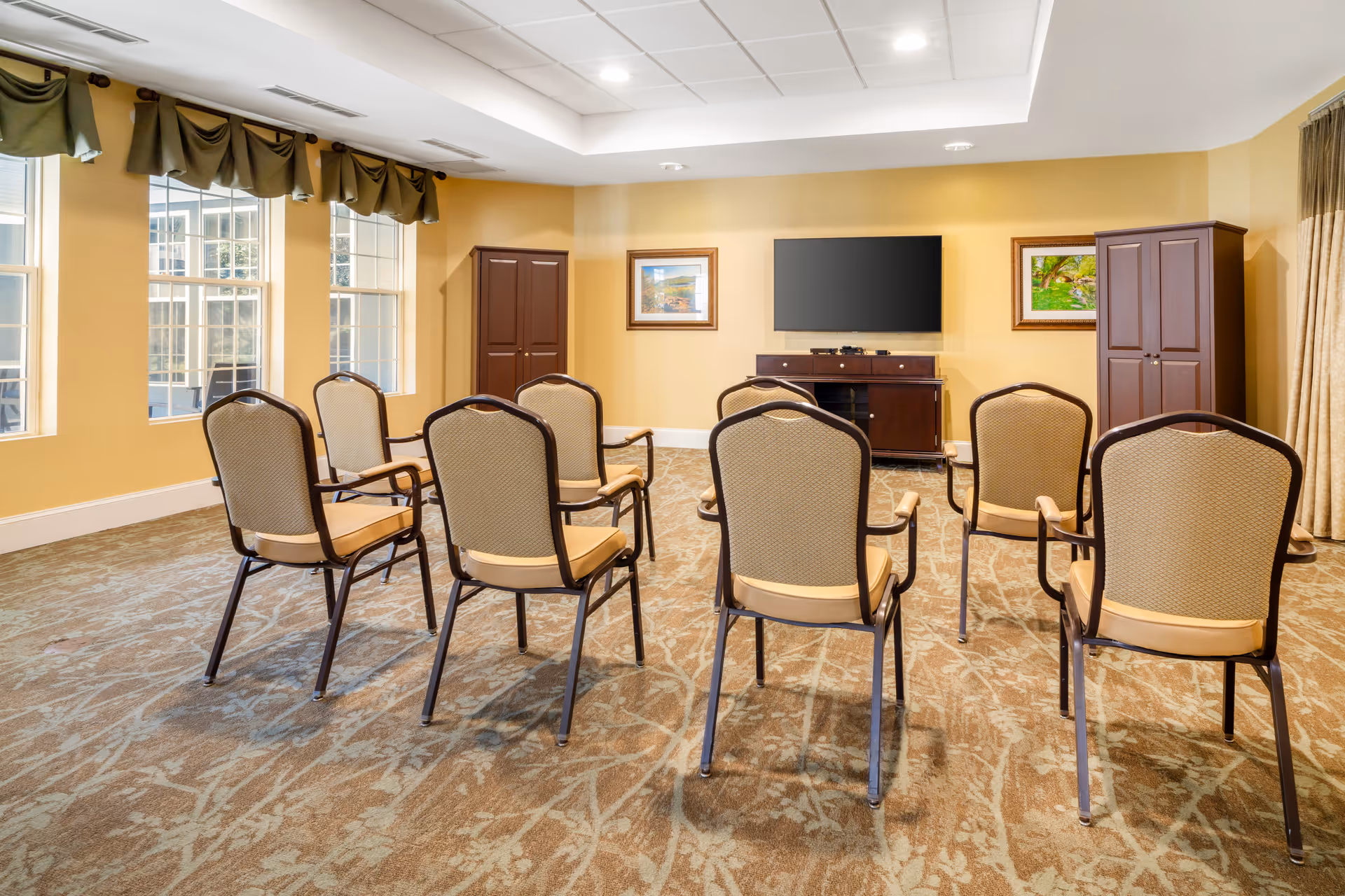 Empty senior living activity room with rows of chairs facing a wall-mounted TV and windows.