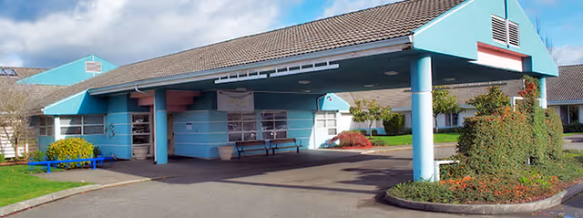 Exterior view of Heartwood Extended Health Care facility showing a covered entrance with blue pillars and a driveway. The building has a tiled roof and surrounding landscaping with bushes and small trees under a partly cloudy sky.