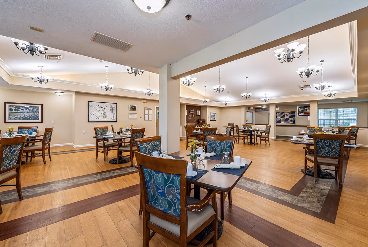 A spacious dining room with multiple wooden tables and chairs arranged neatly. Each table is set with placemats, napkins, cups, and glasses. The room has wooden flooring with decorative inlays and is well-lit with multiple ceiling chandeliers. The walls are adorned with framed pictures and there are windows allowing natural light to enter.