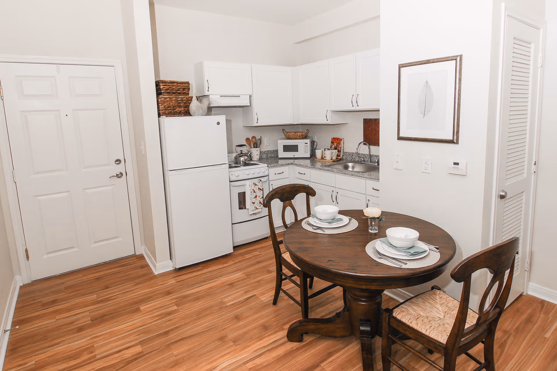 A small kitchen and dining area with white cabinets, a white refrigerator, stove, microwave, and a round wooden table set for two with bowls, plates, and utensils. The floor is wood, and there is a framed leaf artwork on the wall.