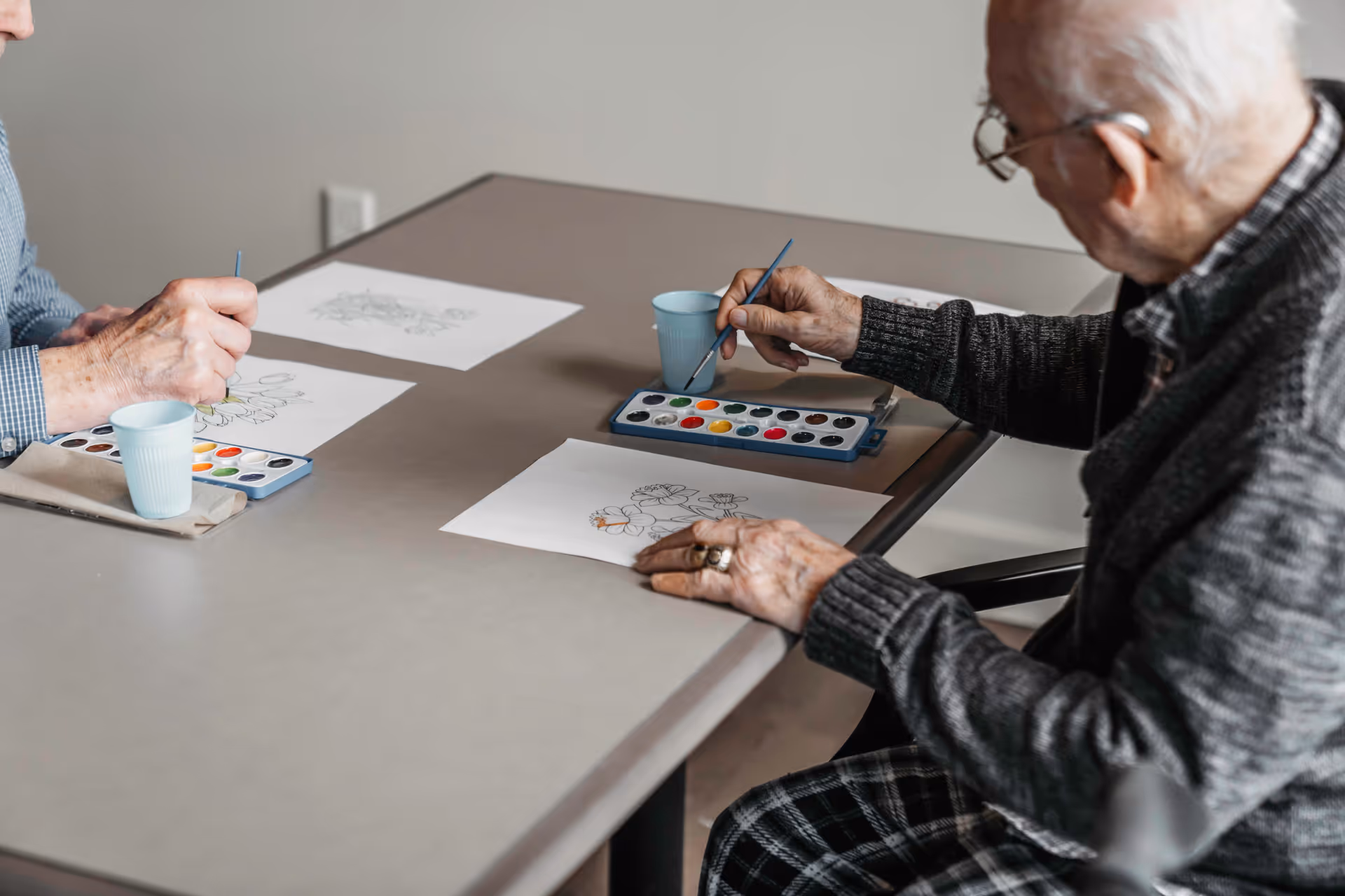 Two elderly individuals seated at a table engaged in watercolor painting, each with a paintbrush and a palette of watercolors, painting floral designs on paper.
