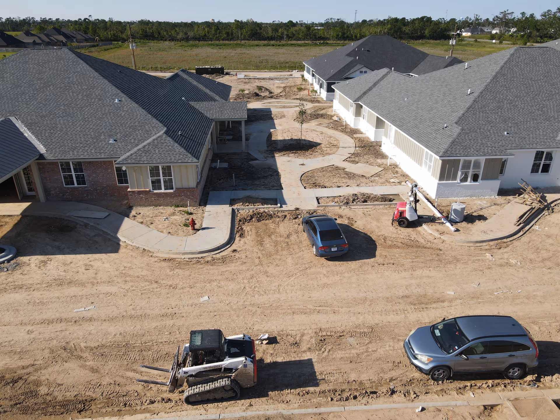 Aerial view of two single-story buildings at a senior living complex under construction with dirt road, parked cars, walkways, and construction equipment.