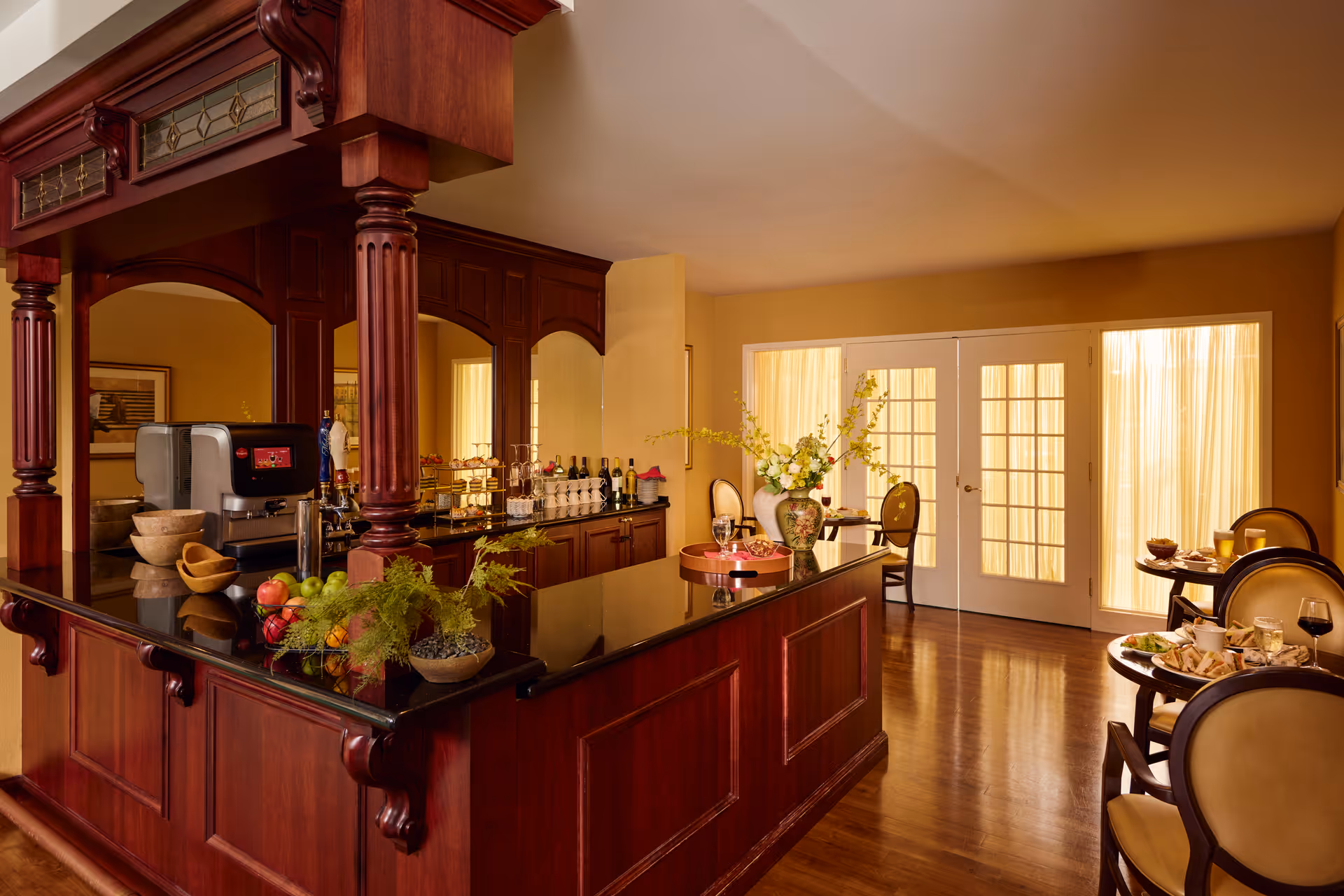 A warm, elegant dining area with a polished wooden bar counter featuring a coffee machine, fruit bowls, and decorative plants. Behind the bar are shelves with bottles and glassware. To the right, there are small round tables set with plates, glasses, and food, with upholstered chairs around them. French doors with sheer curtains allow soft natural light into the room.